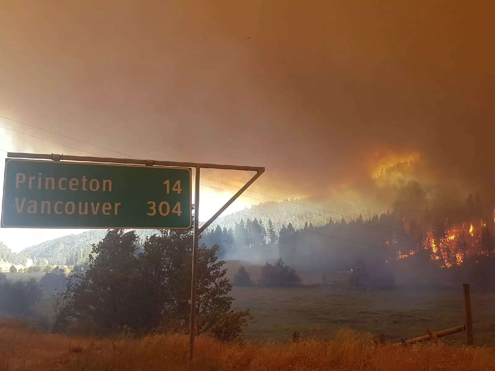 A wildfire burning across a forested hillside beside a rural highway in British Columbia, Canada